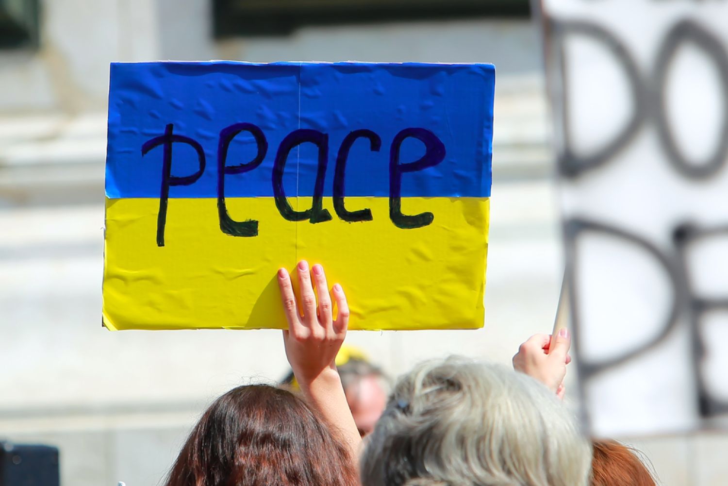 Schild in ukrainischen Nationalfarben mit Aufschrift Peace, von deiner Frau in die Höhe gehalten.